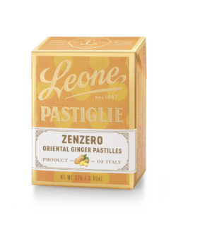 A small, clear tin filled with round, ginger-flavored pastilles. The tin has a brown and white label with the Pastiglie Leone logo and the flavor name in Italian.